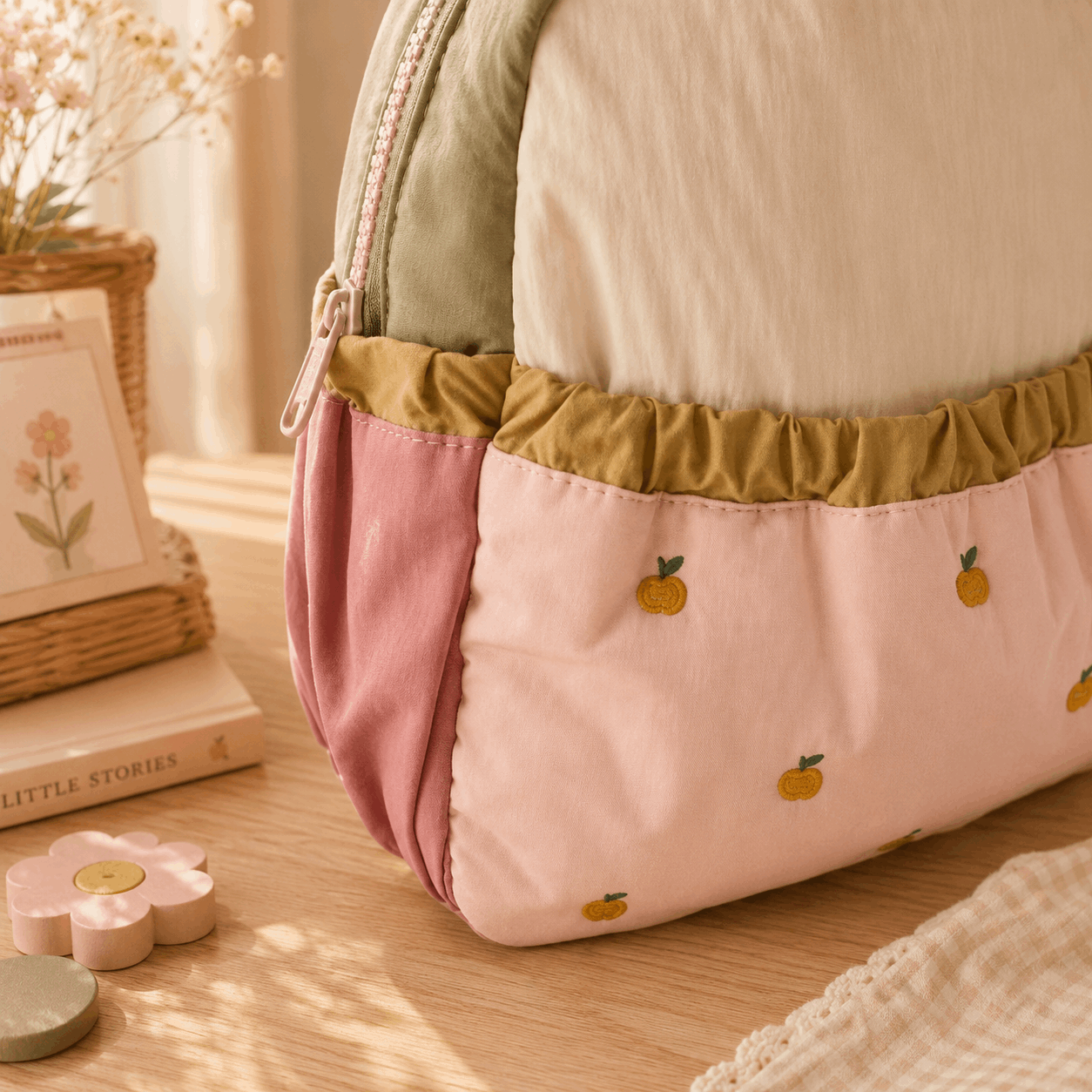 A soft fabric pouch or small bag with a pastel color-blocked design, shown on a wooden surface. The pouch has a cream top section, a dusty pink lower section with small yellow-orange fruit motifs, and a muted olive green ruched band.