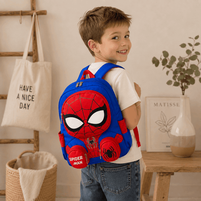 A young boy wearing a white t-shirt and blue jeans is shown from the back, wearing a blue and red Spider-Man themed backpack. The setting is a warm, styled room with a wooden ladder, a canvas tote bag, and a small table with a vase.