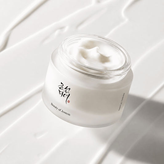 A clear cosmetic jar filled with white cream, photographed on a glossy white surface with diagonal cream streaks and soft shadows. The jar has black Korean lettering and small text reading 'Beauty of Joseon'.