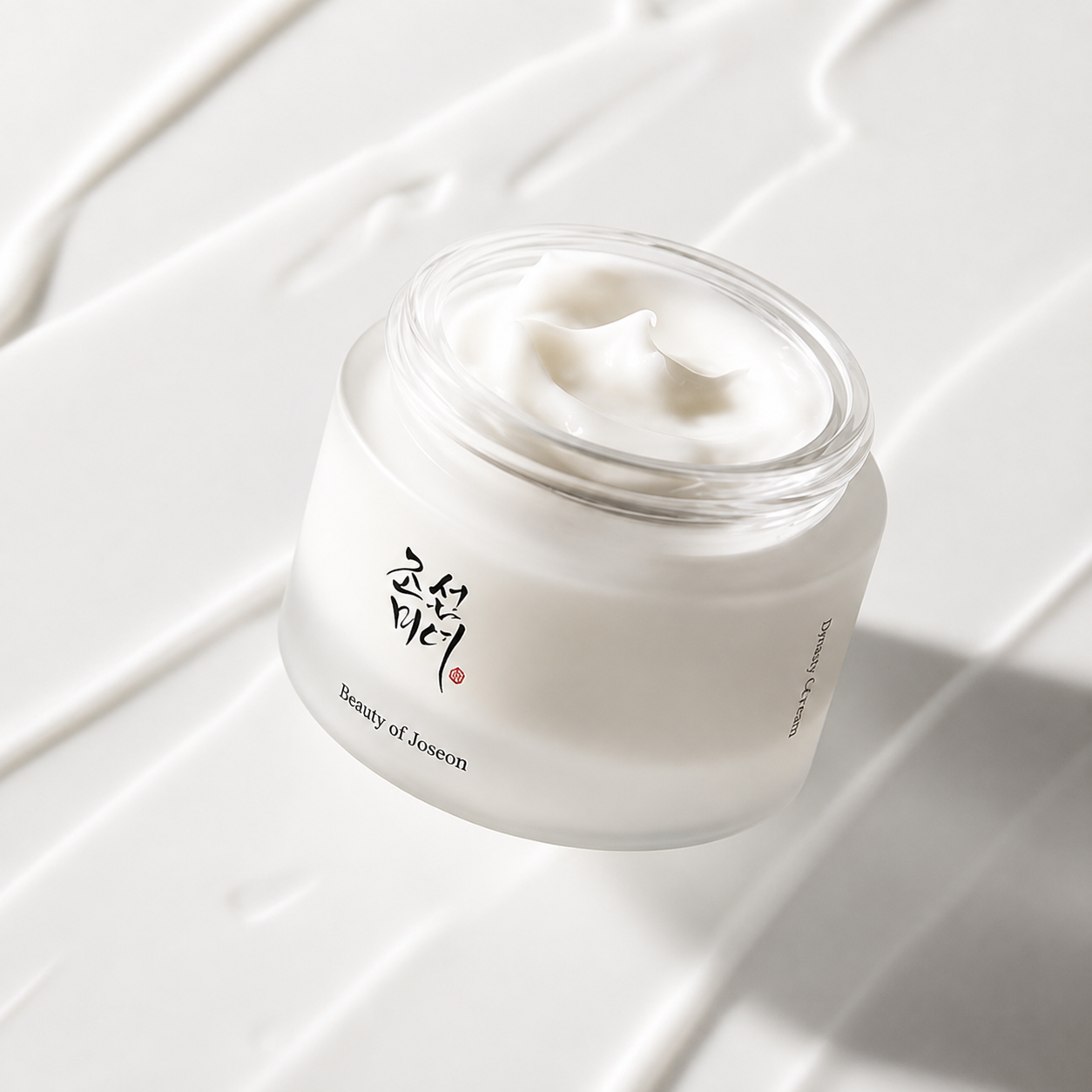 A clear cosmetic jar filled with white cream, photographed on a glossy white surface with diagonal cream streaks and soft shadows. The jar has black Korean lettering and small text reading 'Beauty of Joseon'.