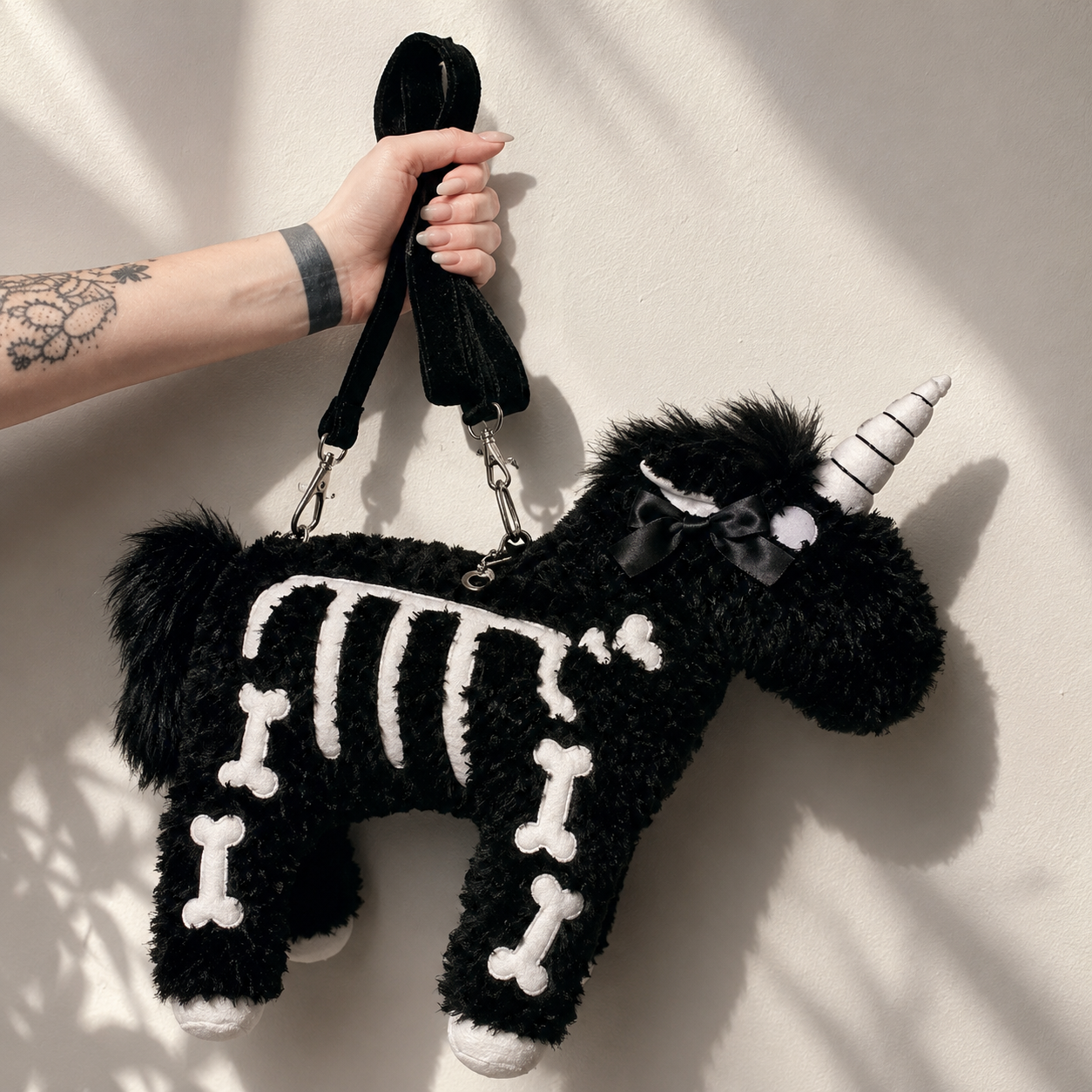 A black plush unicorn-shaped bag with a skeleton bone pattern and a striped horn, held by a tattooed hand against a sunlit beige wall.