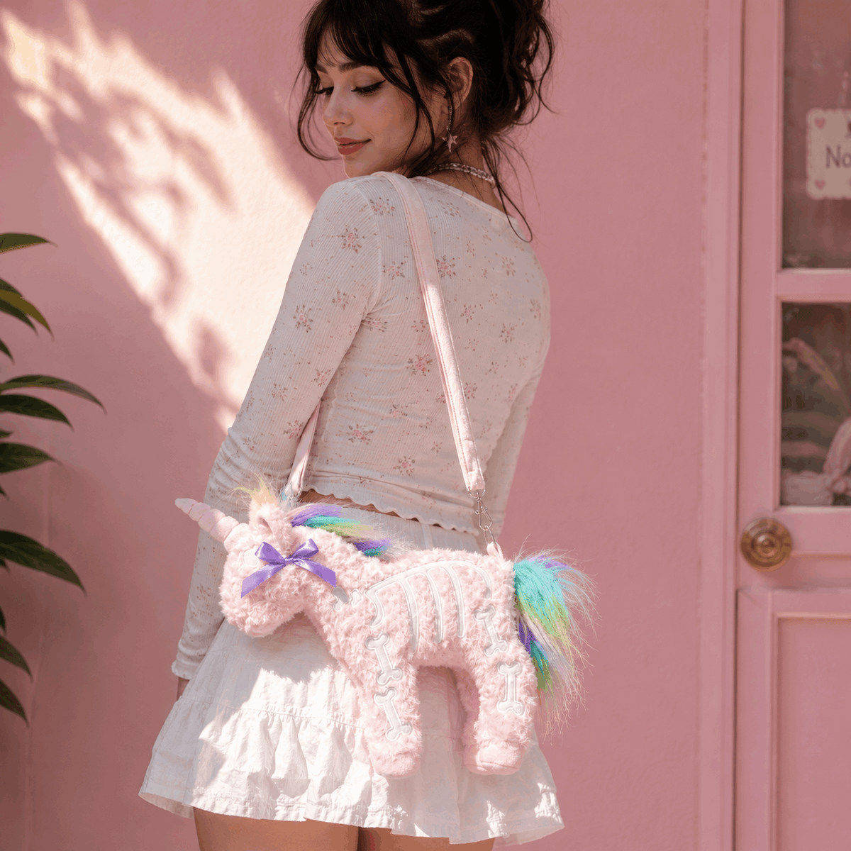 A woman wearing a white long-sleeve floral top and white ruffled skirt carries a pink fuzzy unicorn crossbody bag with a rainbow mane and tail against a pink wall.