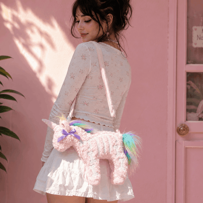 A woman wearing a white long-sleeve floral top and white ruffled skirt carries a pink fuzzy unicorn crossbody bag with a rainbow mane and tail against a pink wall.