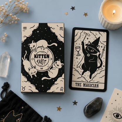 A tarot card deck box and a single card from a 'Kitten Tarot' deck are displayed on a light blue background with mystical props including a lit candle, crystals, dried flowers, and star-shaped decorations. The cards feature whimsical black and cream illustrations of cats in wizard attire.
