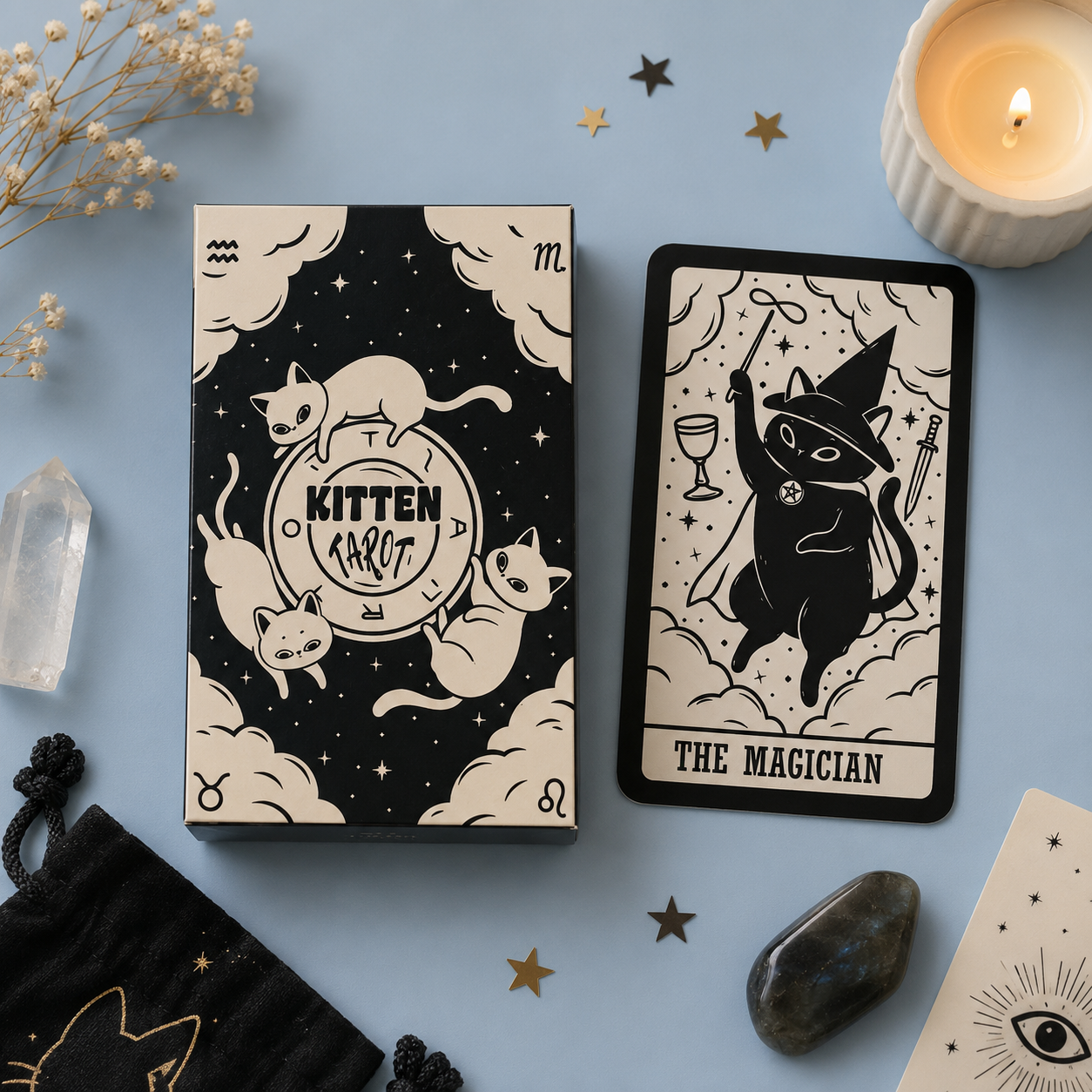 A tarot card deck box and a single card from a 'Kitten Tarot' deck are displayed on a light blue background with mystical props including a lit candle, crystals, dried flowers, and star-shaped decorations. The cards feature whimsical black and cream illustrations of cats in wizard attire.