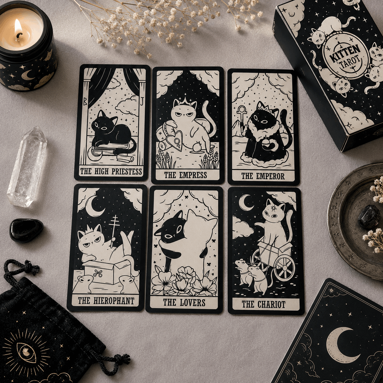 Six illustrated tarot cards featuring cats in black-and-cream artwork are arranged on a light fabric surface, alongside a black box labeled 'Mitten Tarot,' a candle, a crystal, a small pouch, and other mystical props.