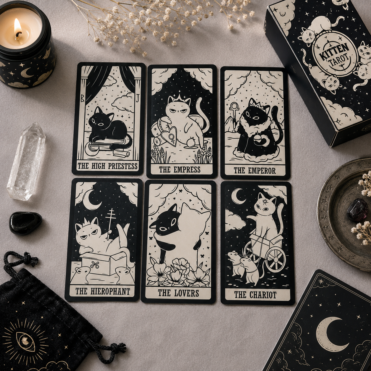 Six illustrated tarot cards featuring cats in black-and-cream artwork are arranged on a light fabric surface, alongside a black box labeled 'Mitten Tarot,' a candle, a crystal, a small pouch, and other mystical props.