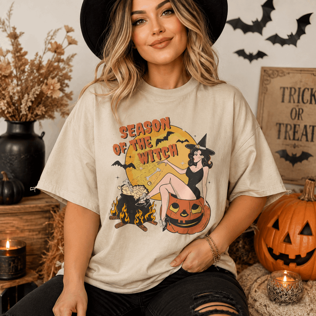 A woman wearing a beige graphic t-shirt with a Halloween design that reads 'SEASON OF THE WITCH' and features a witch, pumpkin, and cauldron. She is styled in a cozy, autumnal setting with pumpkins, candles, and a black hat.