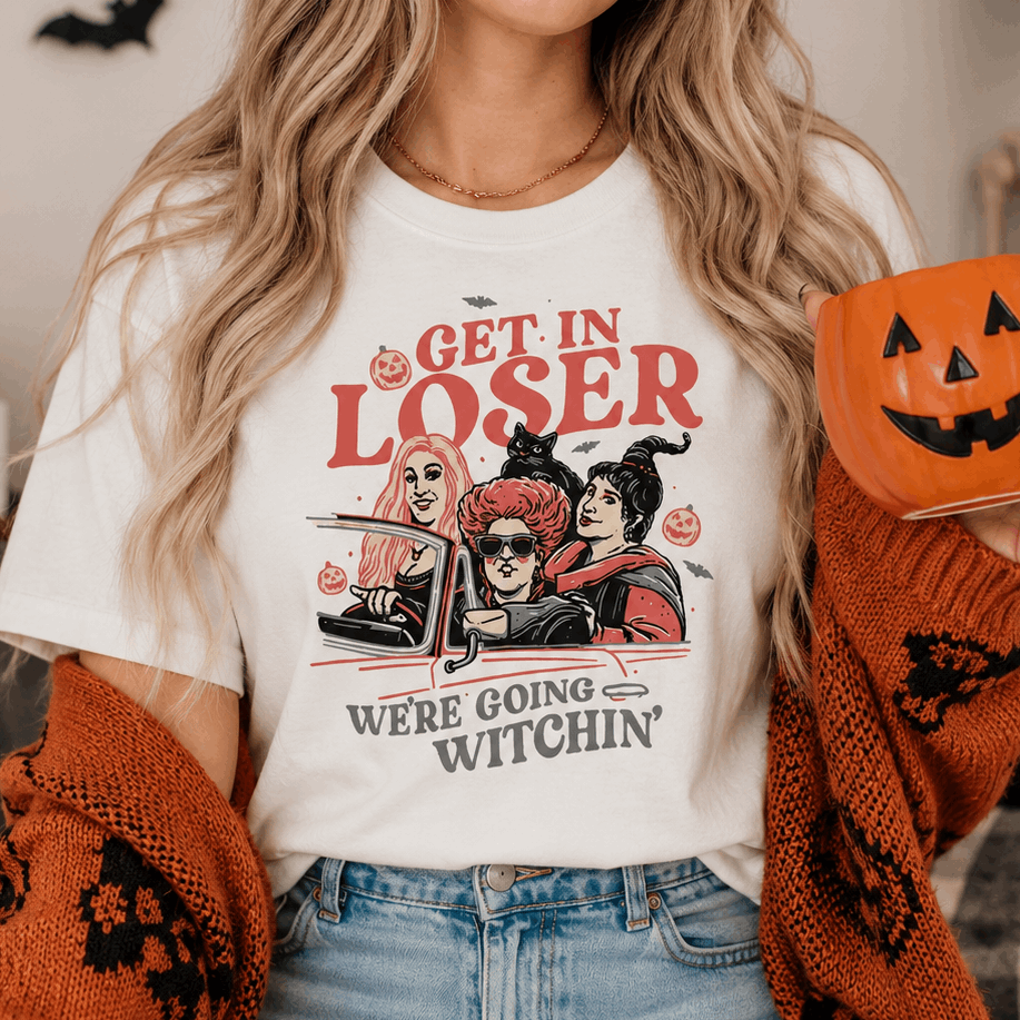 A woman wearing a white graphic t-shirt with a Halloween-themed illustration and text that reads 'GET IN LOSER WE'RE GOING WITCHIN''. She is holding an orange jack-o'-lantern mug and wearing an orange knit cardigan.