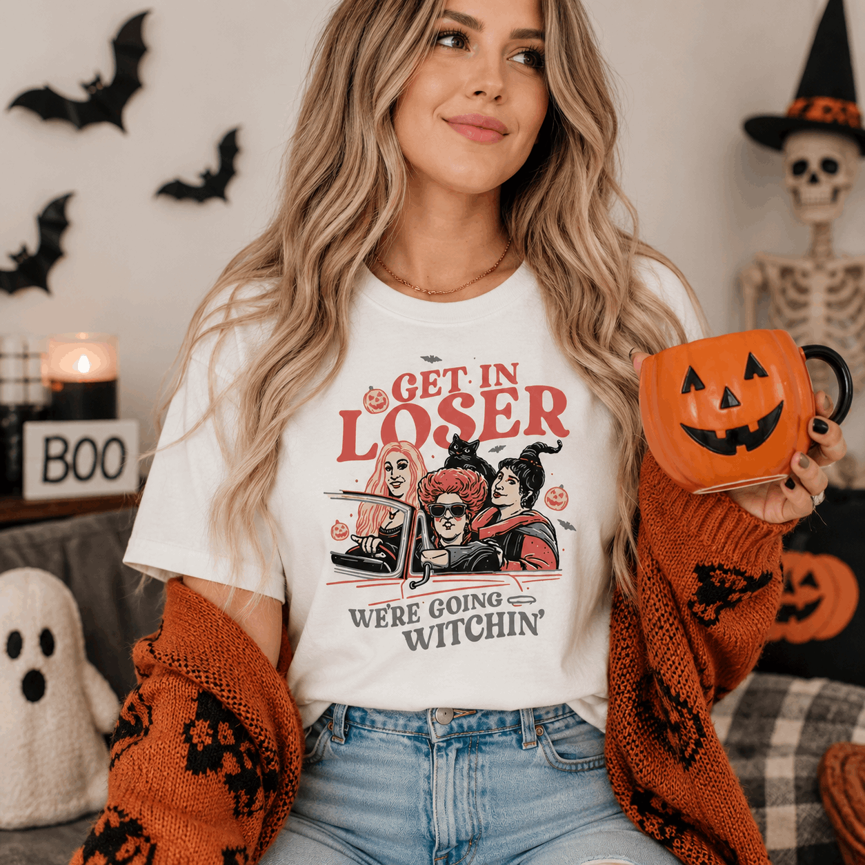A woman wearing a white graphic t-shirt with a Halloween design that reads 'GET IN LOSER WE'RE GOING WITCHIN''. She is holding an orange pumpkin-shaped mug and wearing an orange knit cardigan, set against a cozy, decorated Halloween background with a skeleton, bats, and a ghost.
