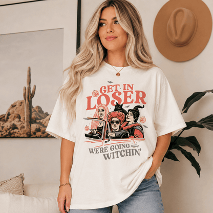 A woman wearing an oversized white graphic T-shirt with a Halloween-themed illustration and the text 'GET IN LOSER WE'RE GOING WITCHIN''. She is standing indoors in a living room setting with a framed cactus print, a hat on the wall, and a plant in the background.