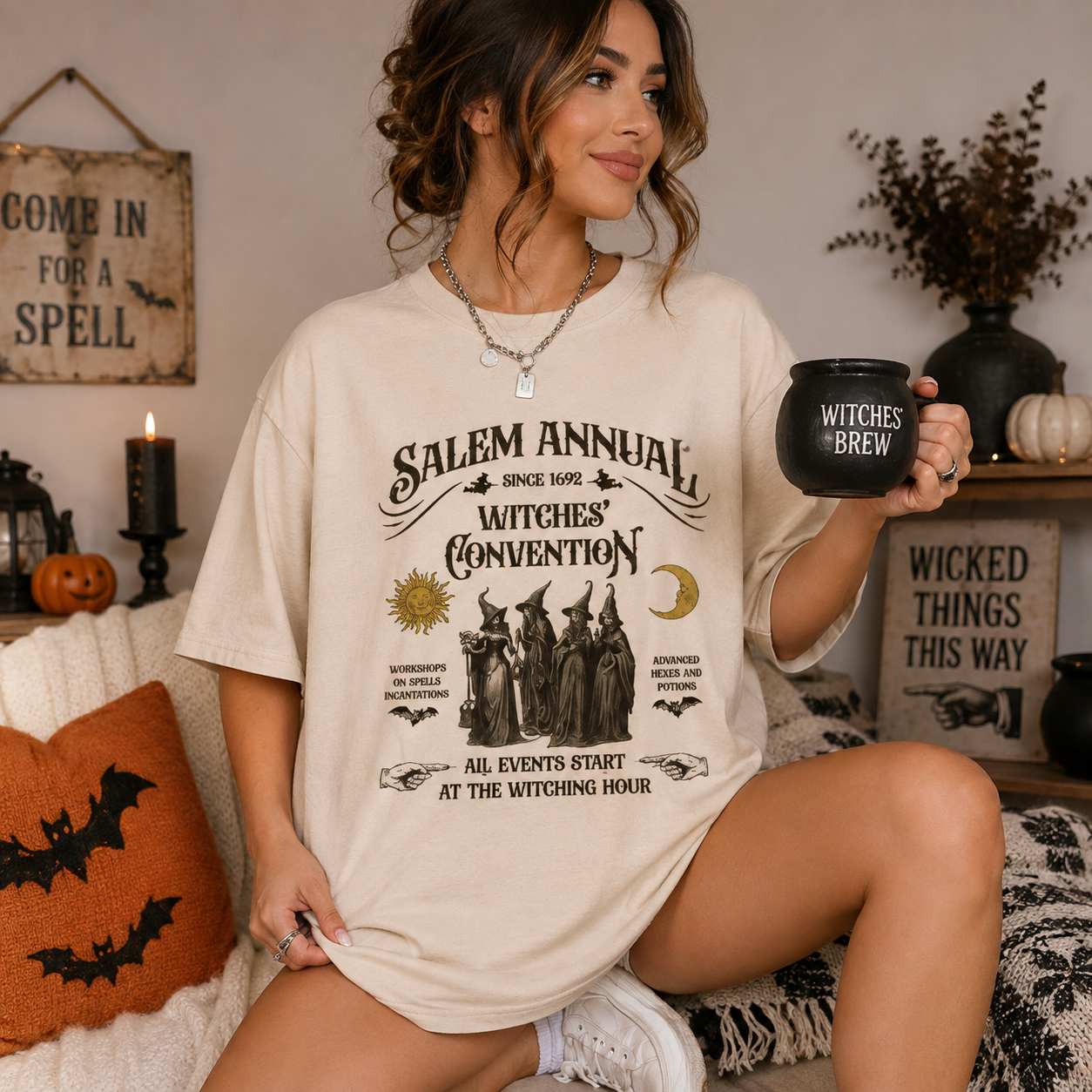 A woman wearing an oversized cream-colored graphic t-shirt with 'Salem Annual Witches' Convention' printed on the front, holding a black mug in a cozy, Halloween-themed room setting.