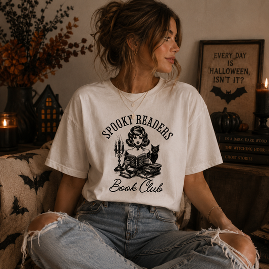 A woman wearing a cream-colored graphic t-shirt with a black print that reads 'SPOOKY READERS Book Club' featuring an illustration of a witch reading under a full moon. She is sitting in a cozy, dimly lit room with Halloween decorations, including a framed sign, candles, and books.