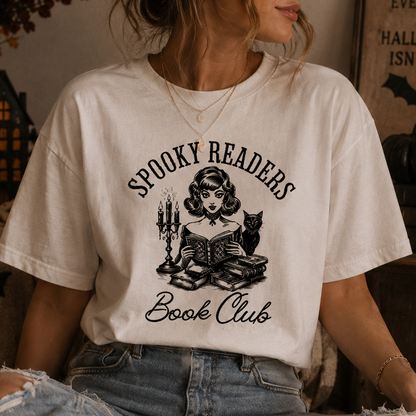 A woman wearing a cream-colored graphic t-shirt with a black illustration of a woman reading, a cat, and candles, with the text 'SPOOKY READERS Book Club'.