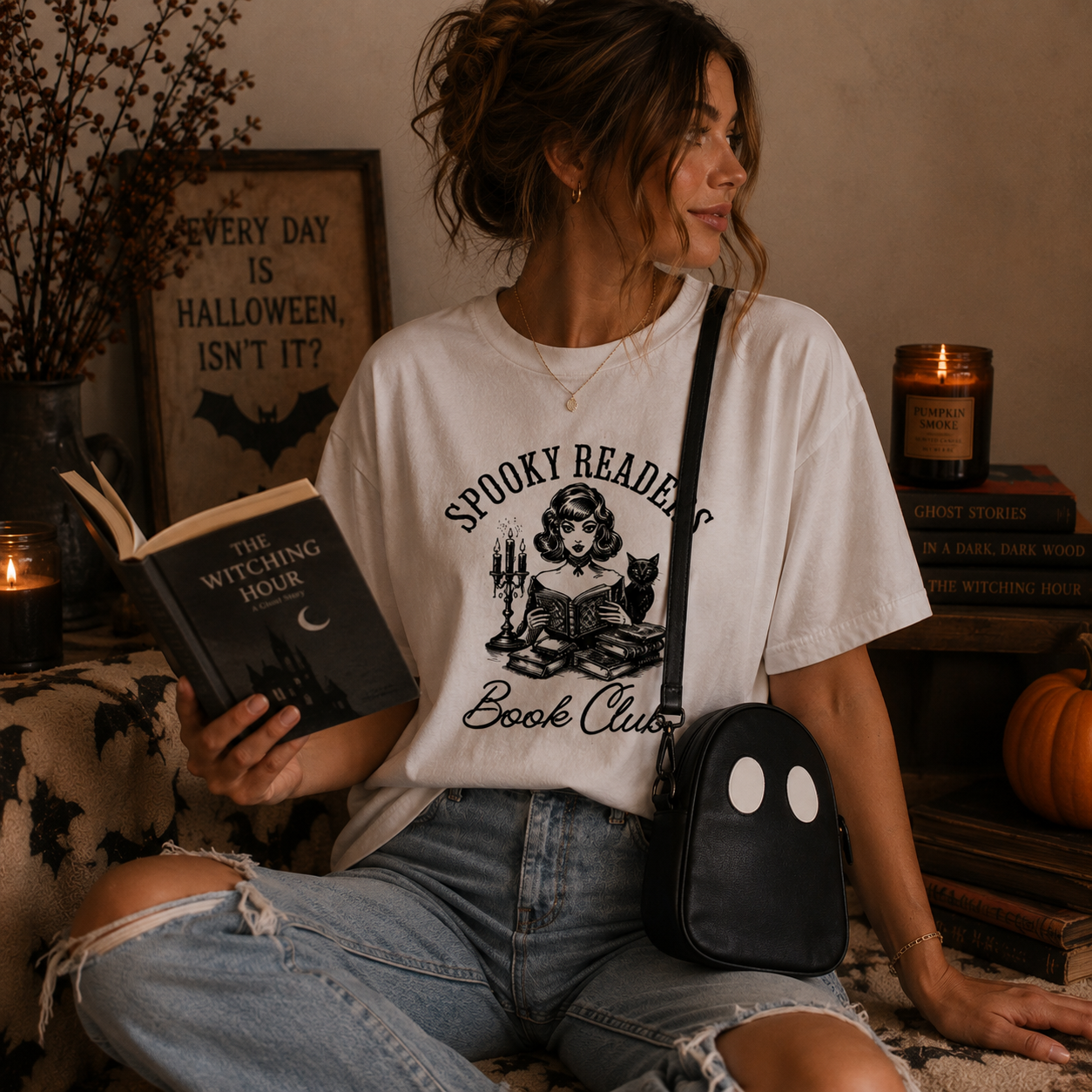 A woman sits in a cozy, Halloween-themed room wearing a white graphic t-shirt that reads 'Spooky Reading Book Club' with a black illustration of a witch. She holds a book and wears a black crossbody bag with a ghost face.