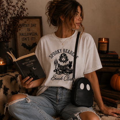 A woman sits in a cozy, Halloween-themed room wearing a white graphic t-shirt that reads 'Spooky Reading Book Club' with a black illustration of a witch. She holds a book and wears a black crossbody bag with a ghost face.