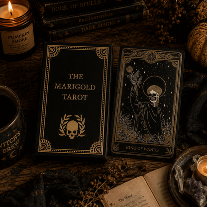 The Marigold Tarot deck displayed with its black and gold box next to a tarot card showing a skull with a halo and raised wand. The scene is styled on a rustic wooden table with candles, dried flowers, an open book, and other occult-themed props.