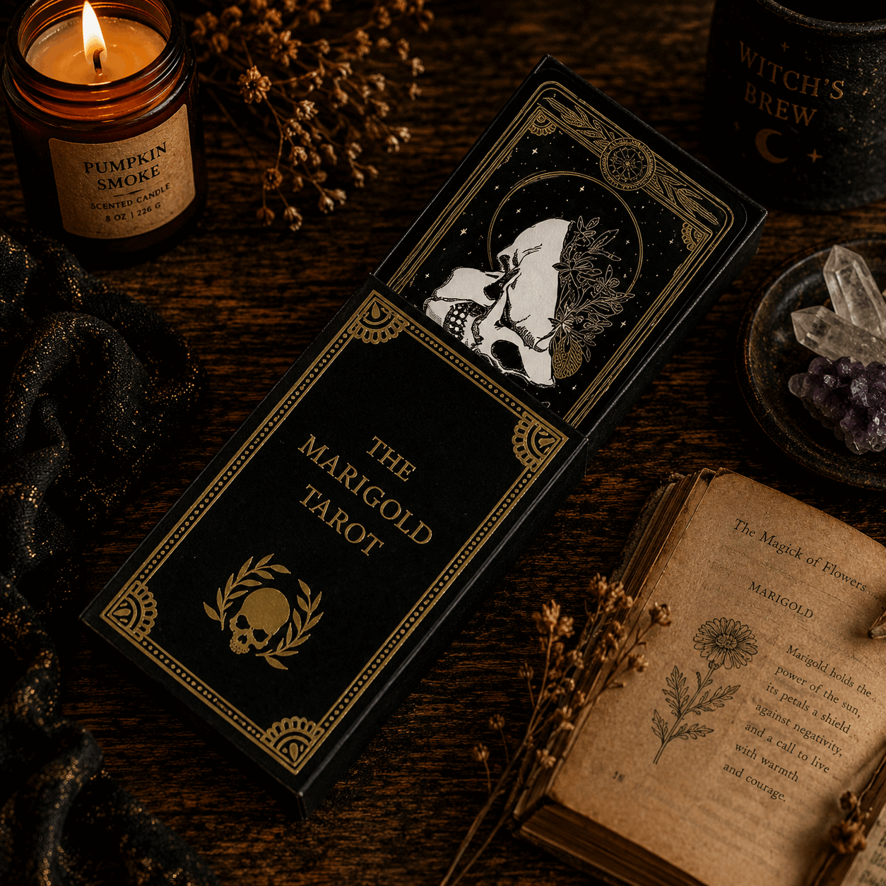The Marigold Tarot deck box, a black tarot card deck in ornate gold-bordered packaging, displayed on a dark wooden table surrounded by mystical props including a lit candle, an open vintage book, crystals, and dried flowers.