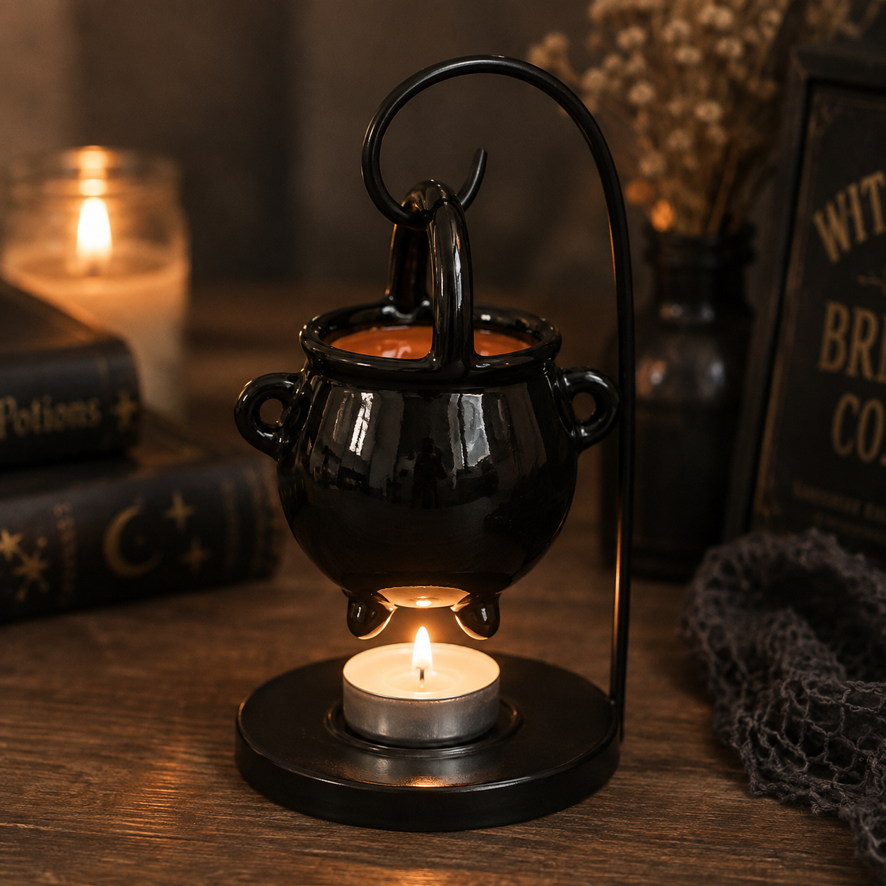 A black cauldron-shaped candle holder with a curved metal stand, holding a tea light candle underneath and a larger orange candle inside, styled on a wooden table with books and a cozy background.