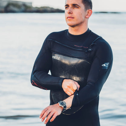 Man wearing a black wetsuit with a brand logo, standing by water.