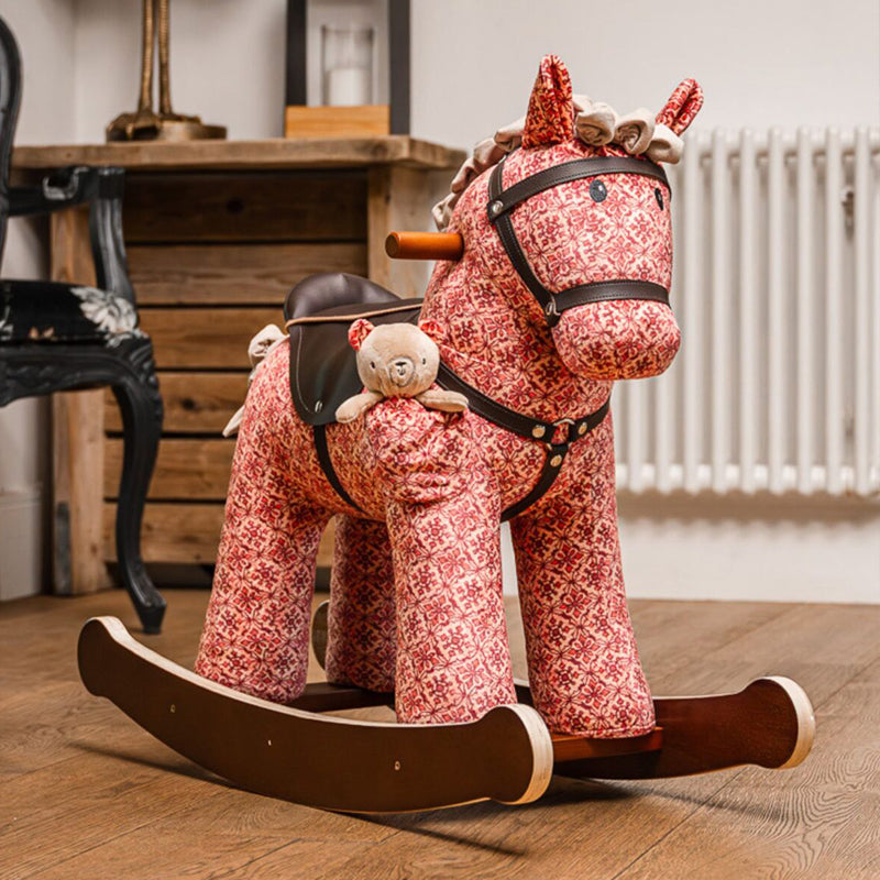 Red floral-patterned rocking horse with a teddy bear on a wooden floor.