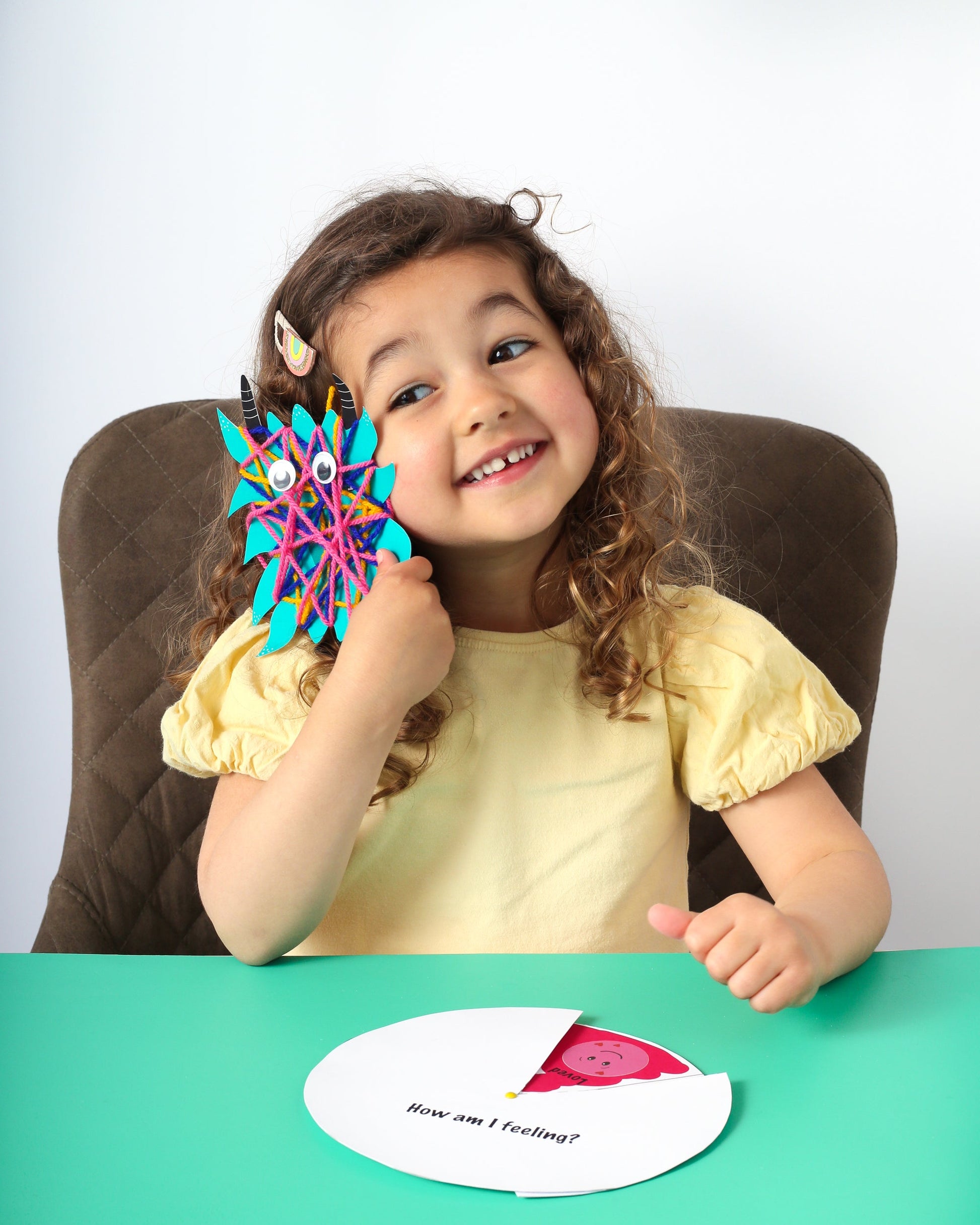 Child holding a colorful toy with googly eyes, sitting at a table with a white card in front of them.