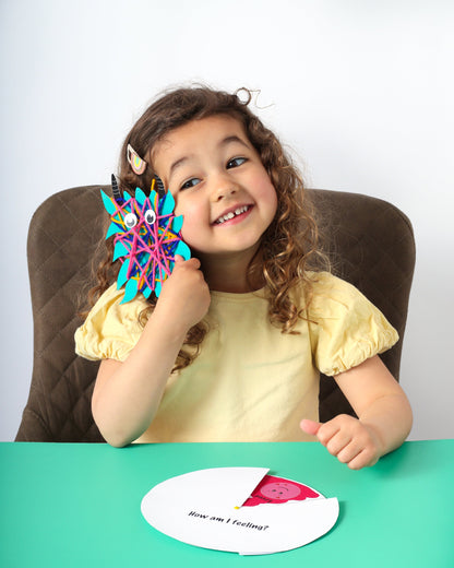 Child holding a colorful toy with googly eyes, sitting at a table with a white card in front of them.