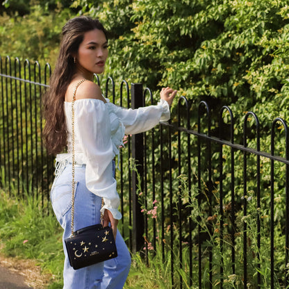 Woman in white blouse and blue jeans holding a black handbag with star designs, standing near a black metal fence.