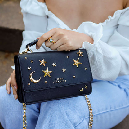 Black handbag with gold star and moon designs held by a person wearing a white top and blue jeans.