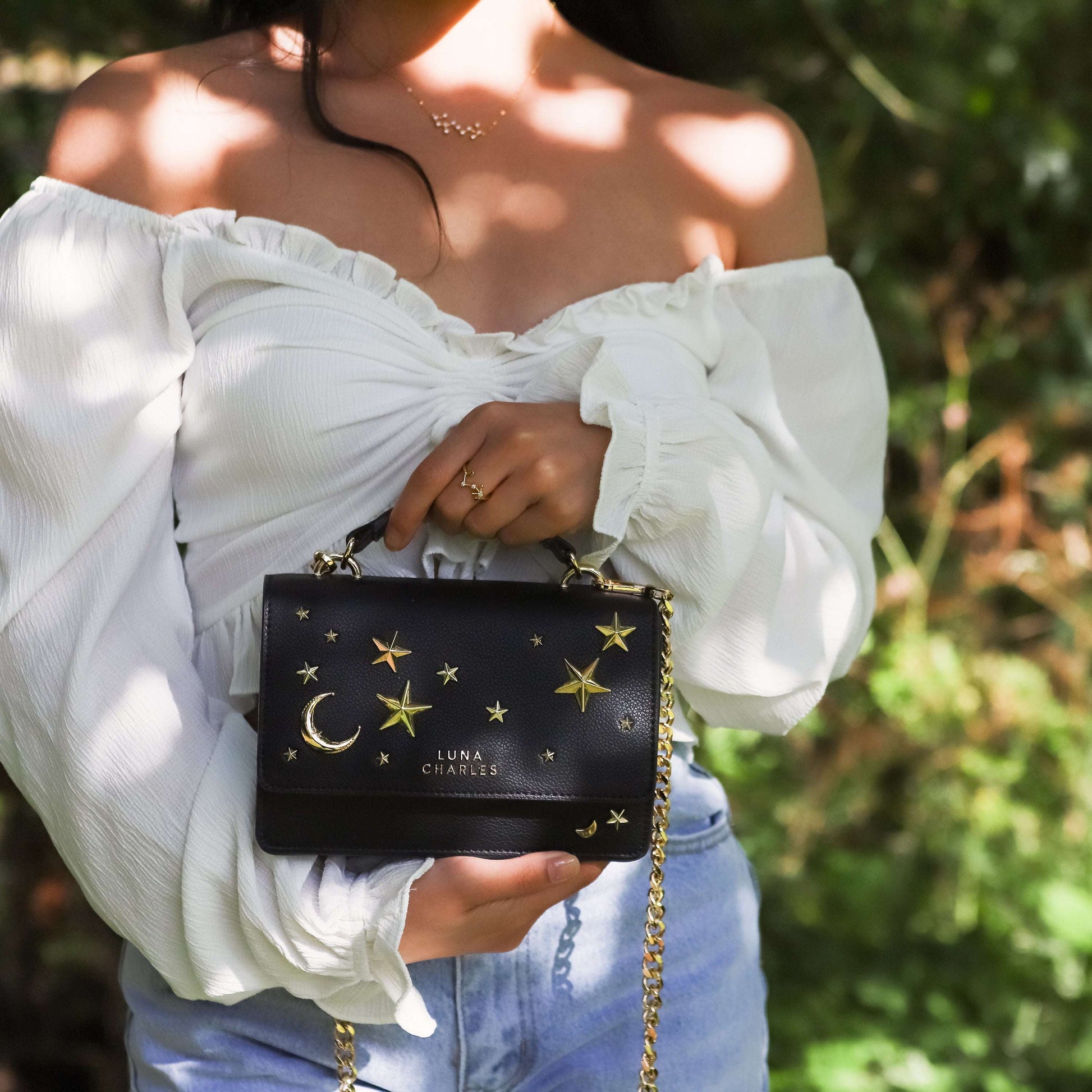 Person holding a black handbag with star and moon designs, wearing a white off-shoulder top and blue jeans.