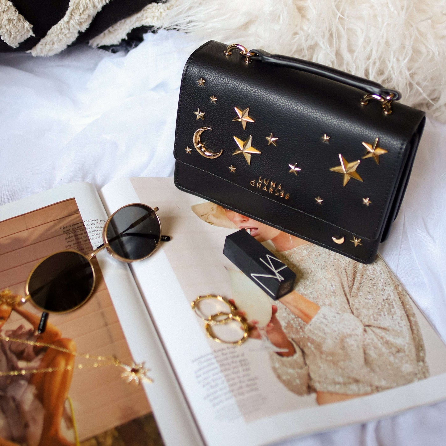 Black handbag with star and moon designs, sunglasses, and a magazine on a white surface
