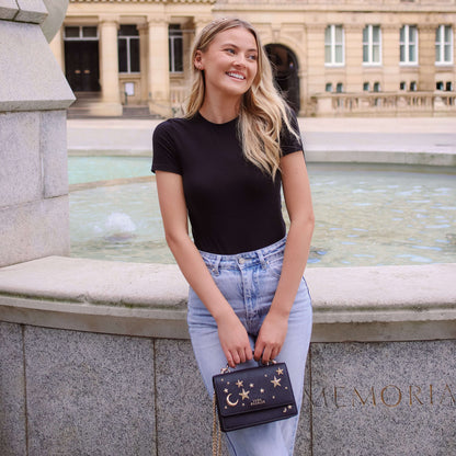 Woman in black top and jeans holding a black handbag with star design, sitting by a fountain.