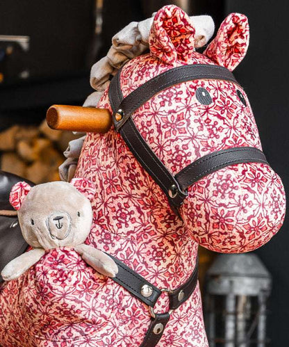 Pink floral-patterned rocking horse with a teddy bear attached, against a blurred background.