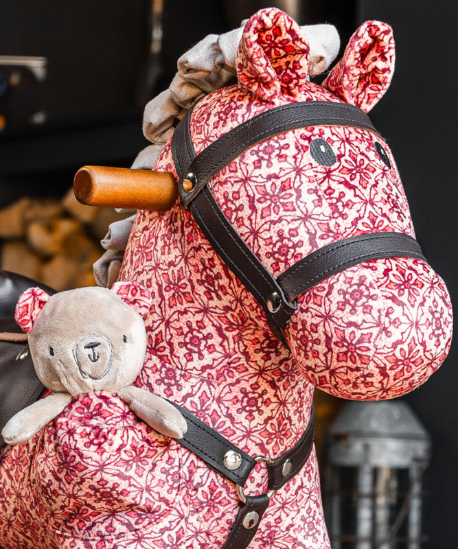 Pink floral-patterned rocking horse with a teddy bear attached, against a blurred background.