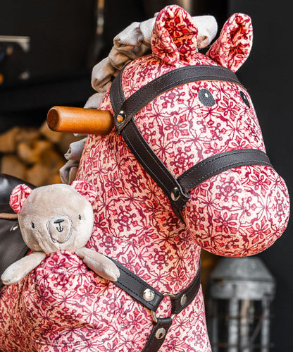 Pink floral-patterned rocking horse with a teddy bear attached, against a blurred background.