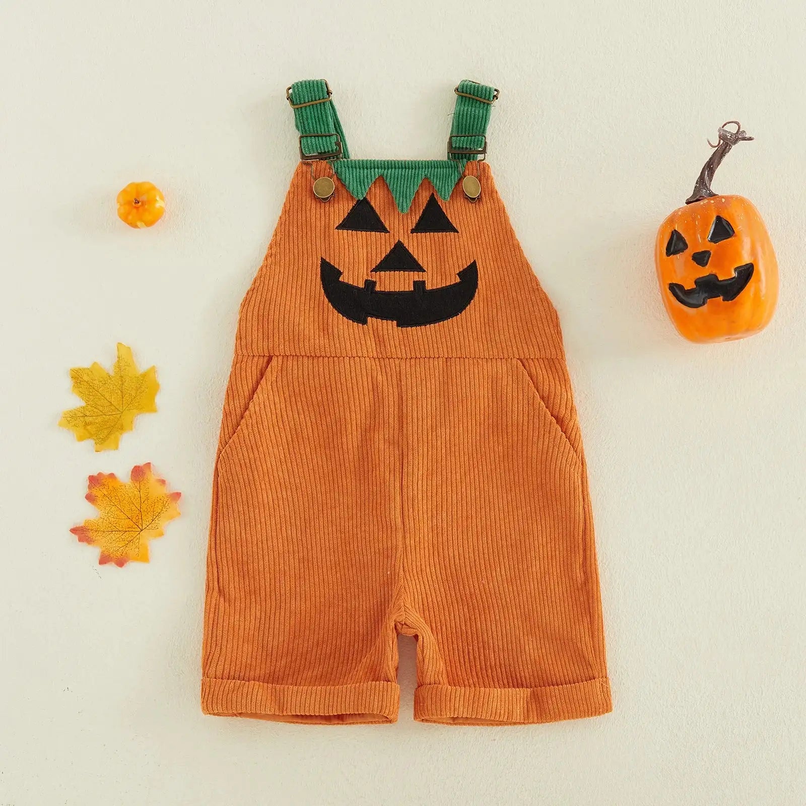 An orange corduroy baby overalls romper styled like a jack-o'-lantern, featuring a black embroidered face and green shoulder straps, displayed on a light cream background with autumn-themed props.