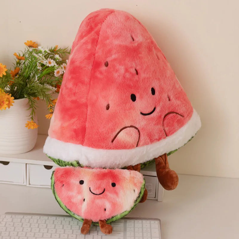 A plush watermelon-themed cushion shaped like a watermelon slice with a smaller matching slice in front, placed on a white desk with flowers and a keyboard in the background.