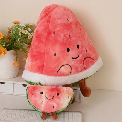 A plush watermelon-themed cushion shaped like a watermelon slice with a smaller matching slice in front, placed on a white desk with flowers and a keyboard in the background.