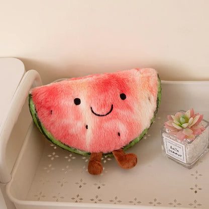 A plush watermelon slice cushion with a smiling face and small brown legs, resting on a white shelf next to a small succulent in a glass jar.