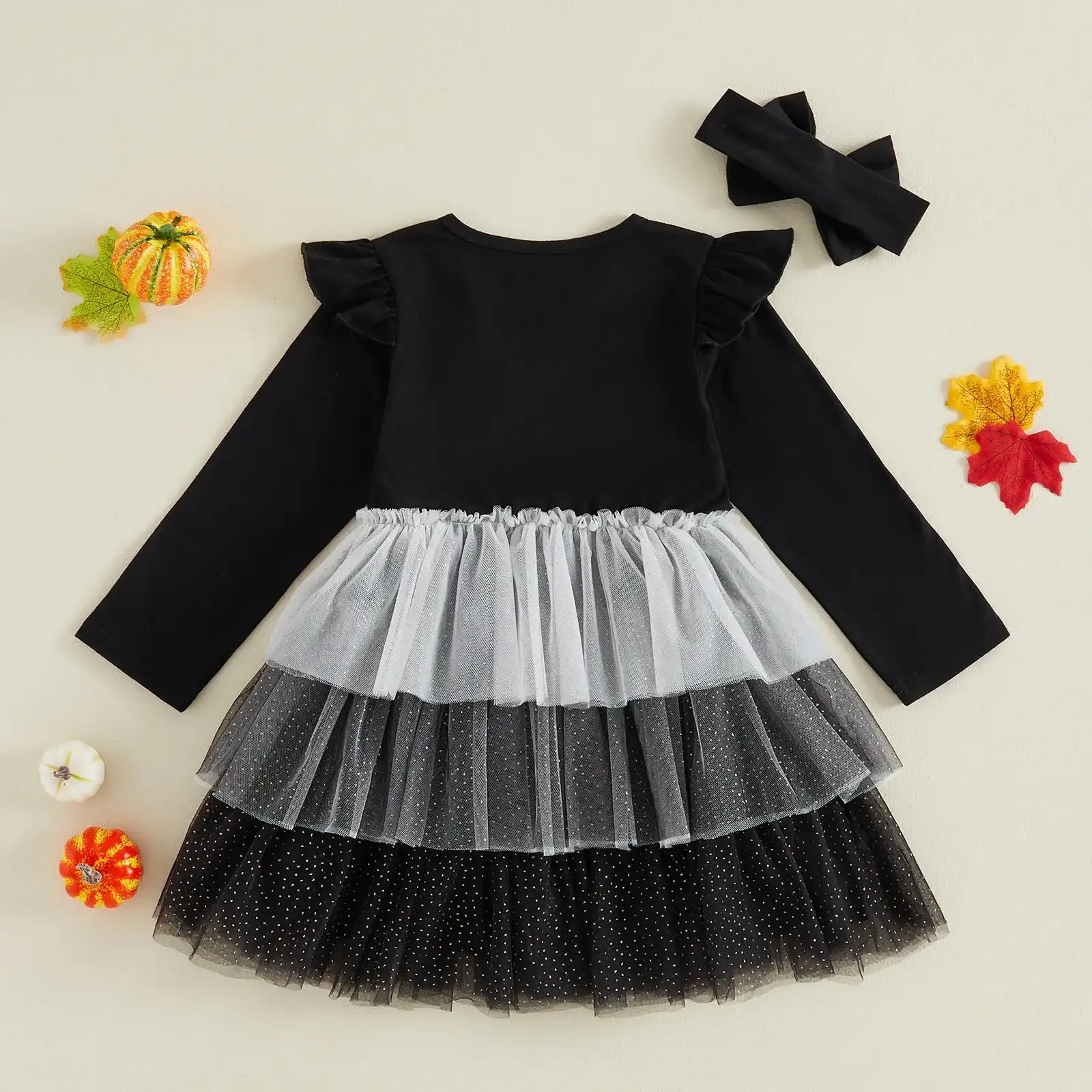 A toddler girl's Halloween costume set featuring a long-sleeve dress with a black top, white and black tiered tulle skirt, and a matching black bow headband, displayed on a beige background with decorative autumn props.