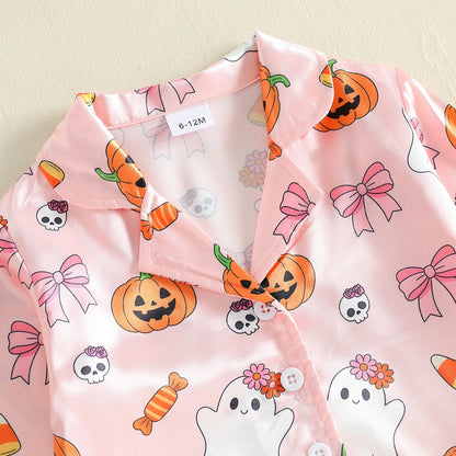 A close-up of a light pink pajama shirt with a Halloween print featuring orange jack-o'-lanterns, pink bows, white skulls, and a white ghost with flowers, laid flat on a light beige background.