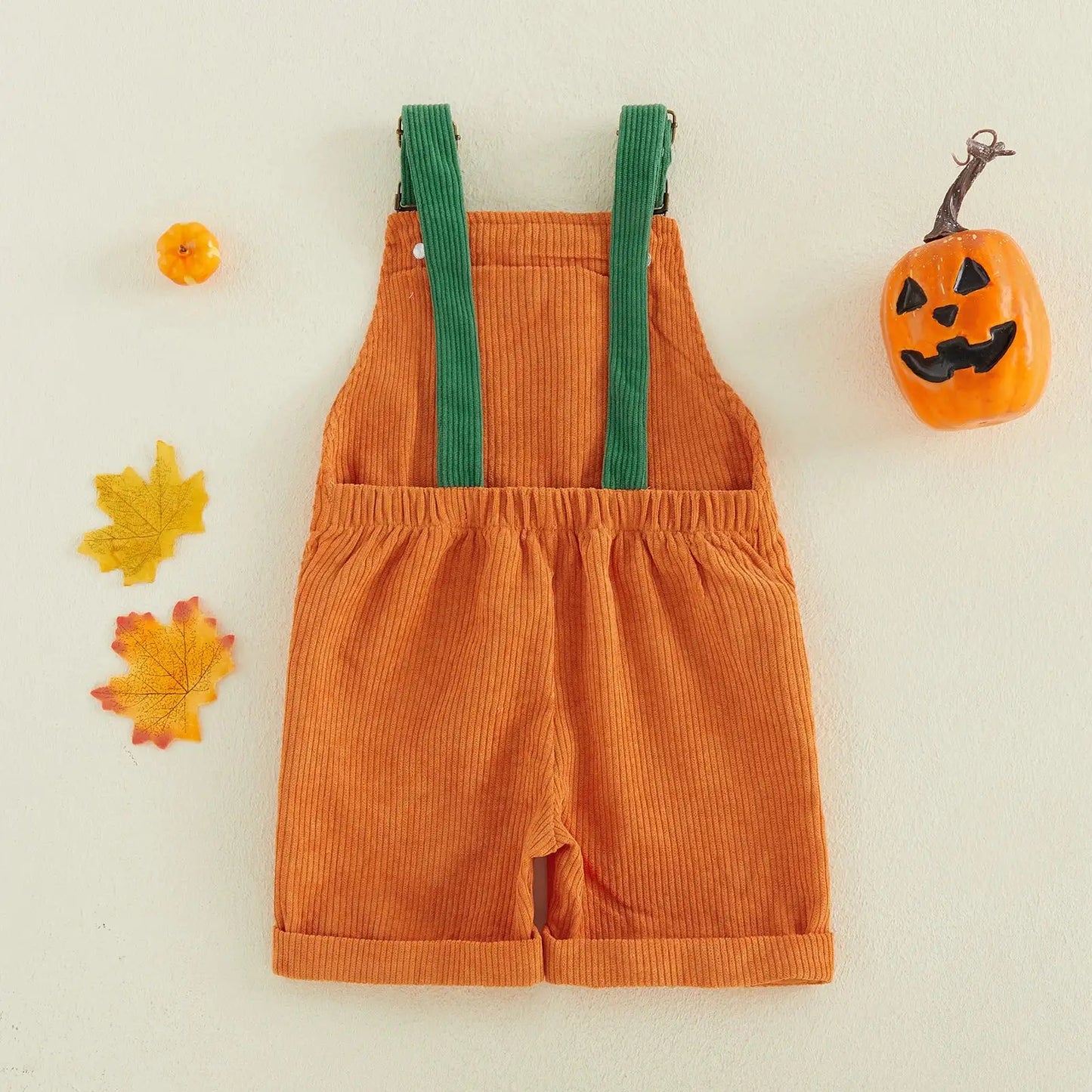 An orange corduroy shortall romper with green shoulder straps, styled flat on a light cream background with autumn props including a jack-o'-lantern, maple leaves, and a small pumpkin.