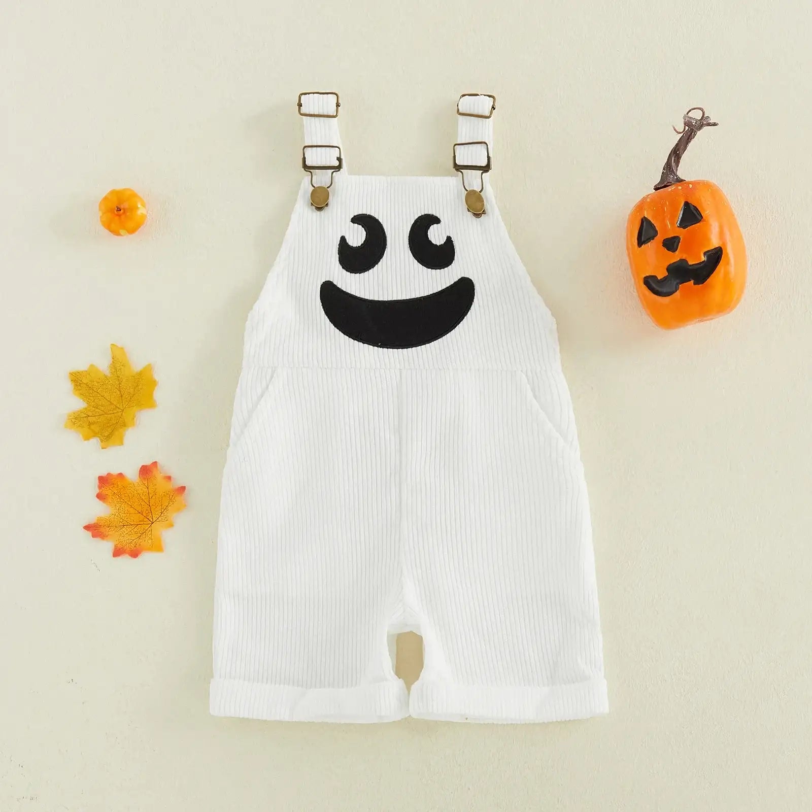 White baby overalls with a black ghost face graphic on the chest, styled flat-lay with small orange pumpkins and autumn leaves on a cream background.