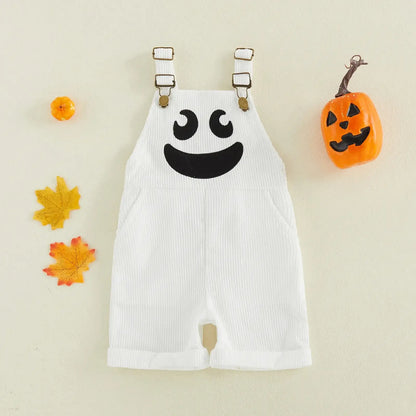 White baby overalls with a black ghost face graphic on the chest, styled flat-lay with small orange pumpkins and autumn leaves on a cream background.