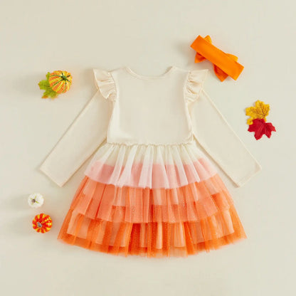 A baby girl's long-sleeve dress with a cream top and a tiered tulle skirt in white, pink, and orange, styled flat-lay with autumn props on a light beige background.