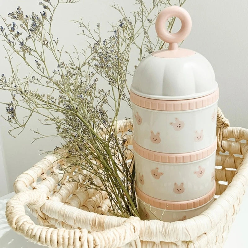 Three-tiered cylindrical container with pink accents and animal prints on a woven tray with dried plants.
