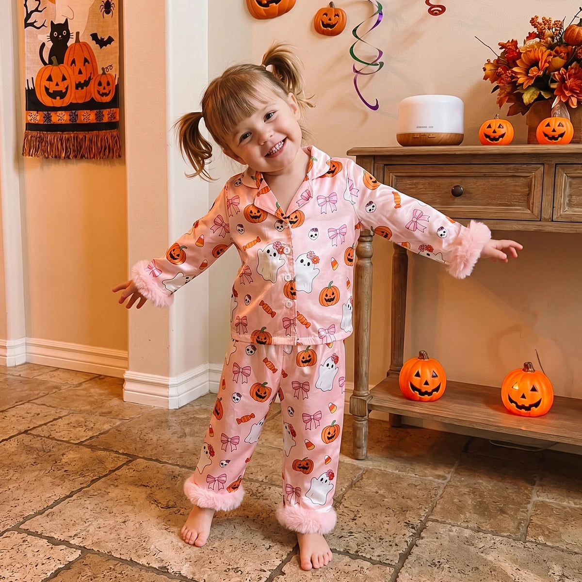 A young girl with blonde pigtails stands barefoot in a decorated indoor room wearing a pink Halloween-themed pajama set with a ghost and pumpkin print and fluffy trim. She is posed in front of a wooden console table with jack-o'-lanterns and autumn decorations.
