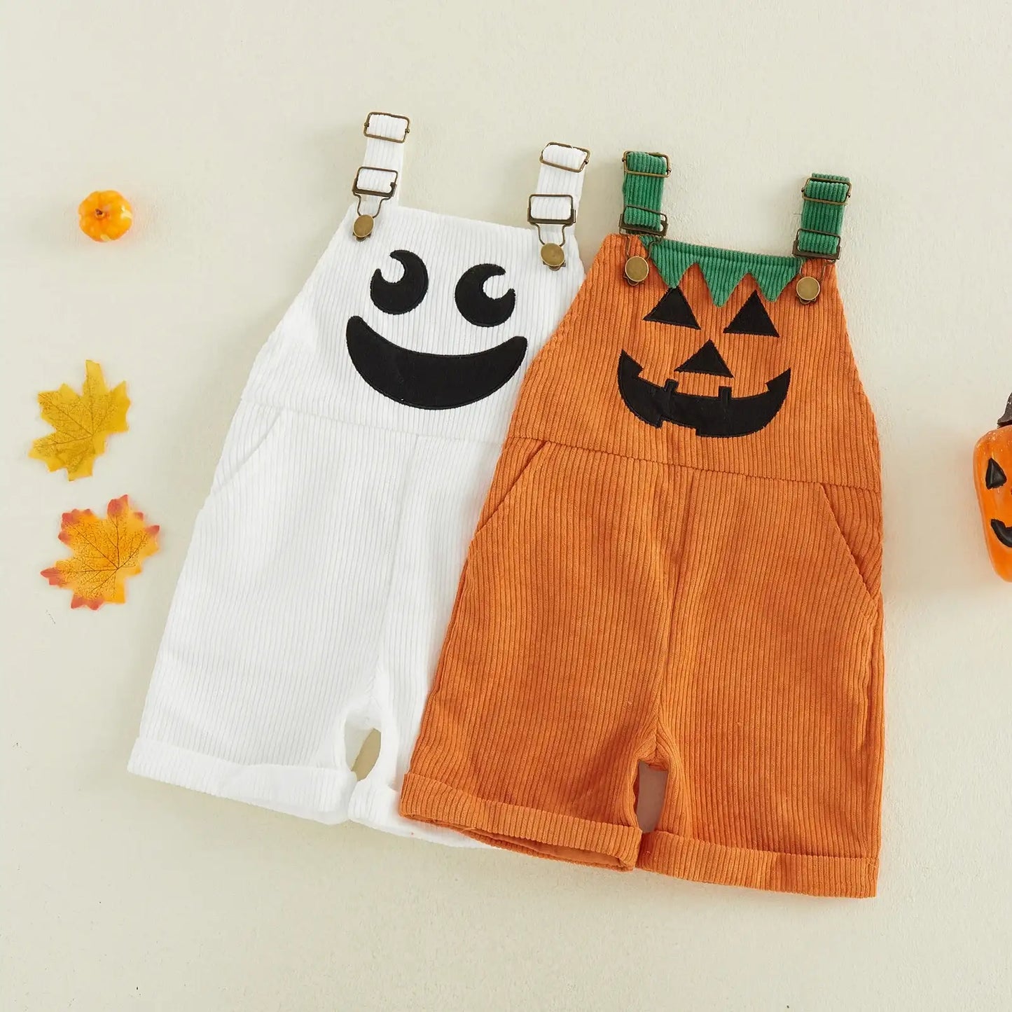 Two children's Halloween corduroy overalls laid flat on a light cream background. One is white with a black ghost face and silver buckle straps, and the other is orange with a black jack-o'-lantern face and green shoulder straps.