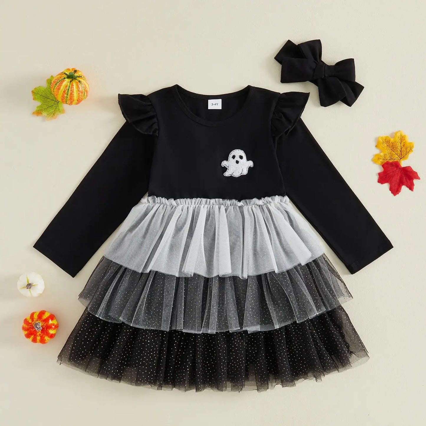 A toddler girl's Halloween dress with a black long-sleeve top, a white ghost applique on the chest, and a tiered tulle skirt in black, gray, and white. A matching black bow headband is placed above the dress, and small autumn leaves and pumpkins decorate the background on a light cream surface.
