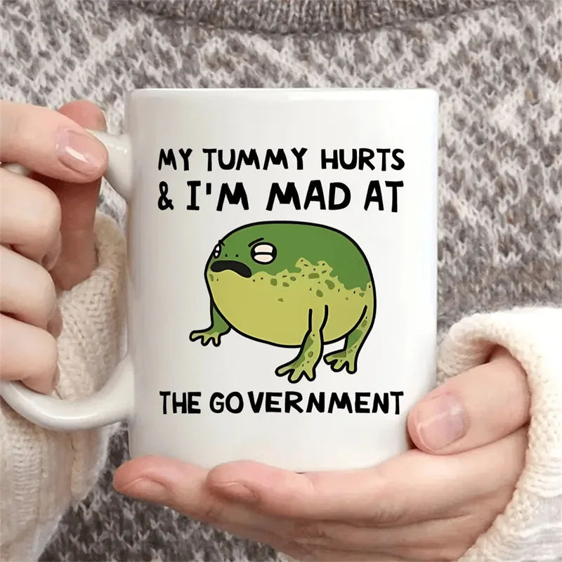 Mug with a green frog illustration and text held by hands against a textured background